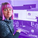 Fanny mit Schnute vor ihrem Monitor an ihrem Arbeitsplatz (lila eingefärbt mit Tastatur, Maus und Mikro). Auf dem Monitor ist die Webseite und das Instagram-Profil zu sehen.